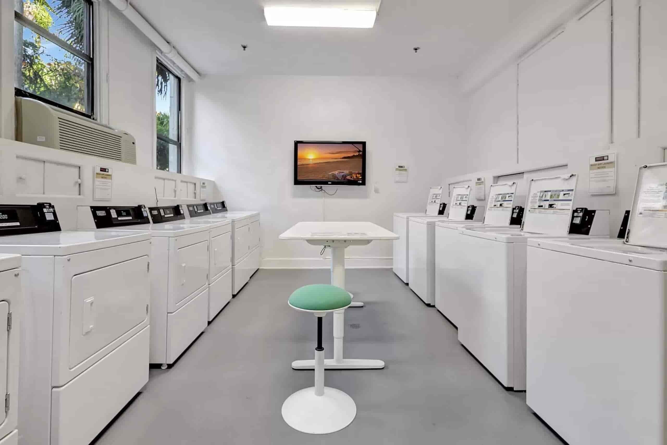 Eine Wäscherei mit Reihen von Waschmaschinen, einem Tisch in der Mitte mit einem Stuhl und einem an der Wand befestigten Fernseher.