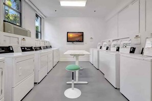 Een wasserette met rijen wasmachines, een centrale tafel met een stoel, en een televisie aan de muur.