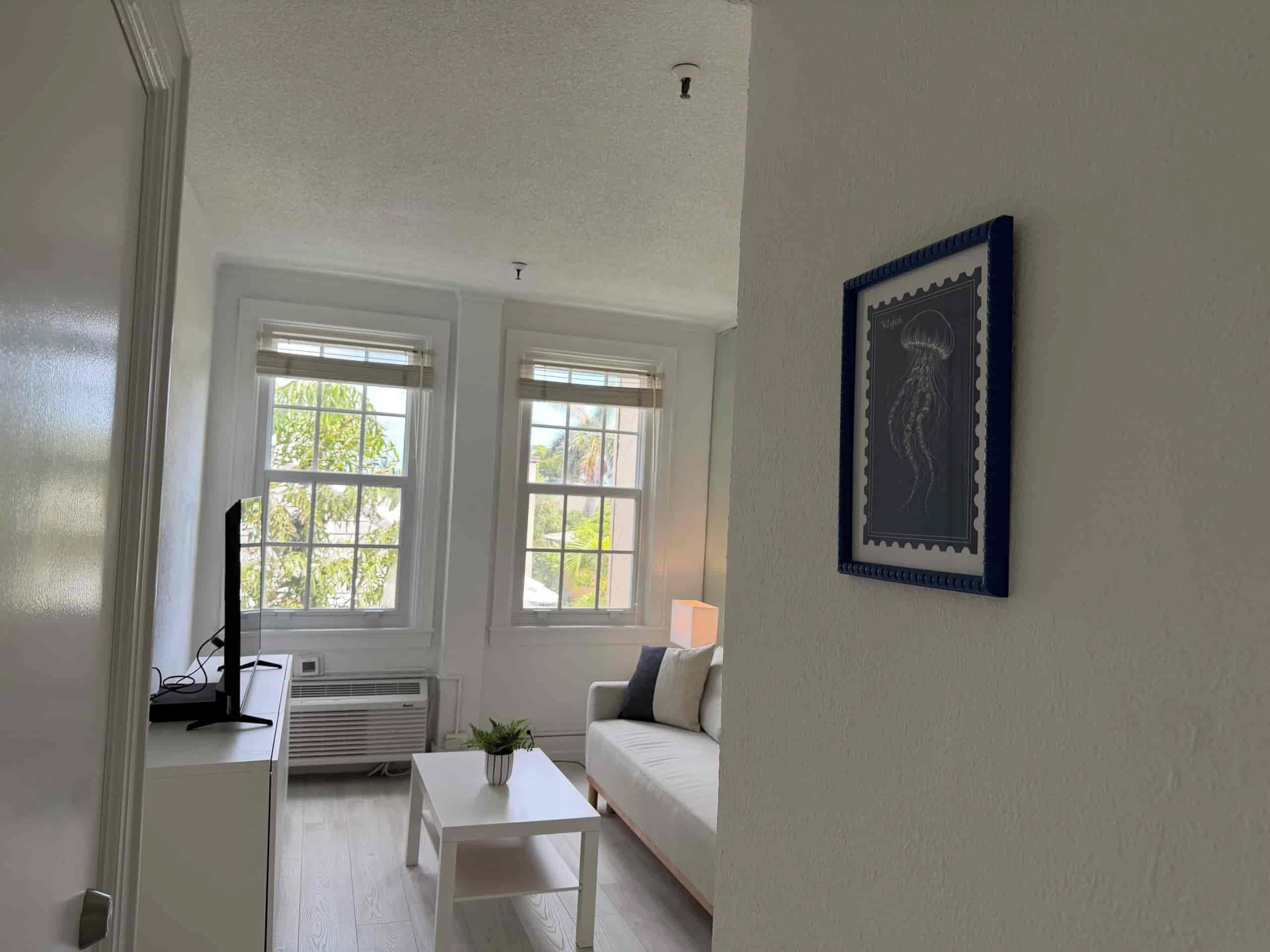 Ein Zimmer mit einem weißen Sofa, einem kleinen weißen Tisch und zwei Fenstern mit weißen Rahmen.