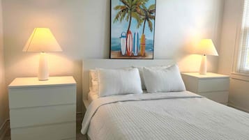 A bedroom with a bed, two bedside tables, a painting of surfboards, and a window with blinds.