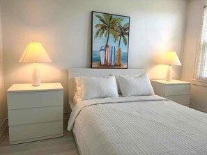 Un dormitorio con cama, dos mesitas de noche, un cuadro de tablas de surf y una ventana con persianas.