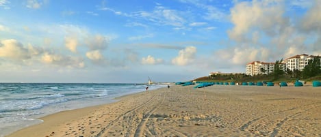 A beach with footprints leading to the water, a building with multiple balconies, and a clear sky.