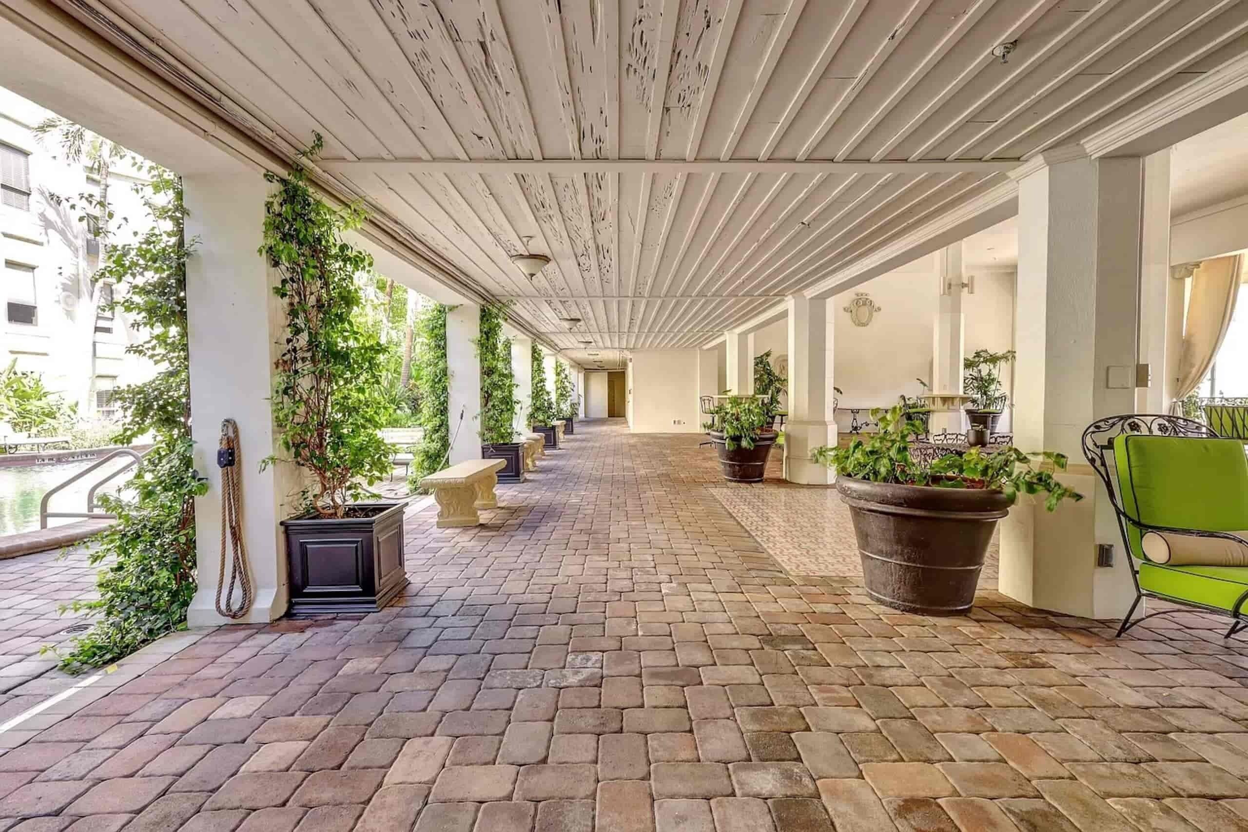 A covered walkway with potted plants and benches, leading to a building entrance.