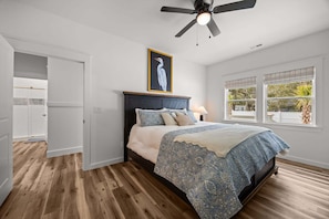 A bedroom with a bed, a ceiling fan, a window with a view, and a door leading to a bathroom. - Holy Crab!: 4BR 4BA, Pool, Fire Pit, EV Charger, Close to Beach, Sleeps 14 (Oak Island)