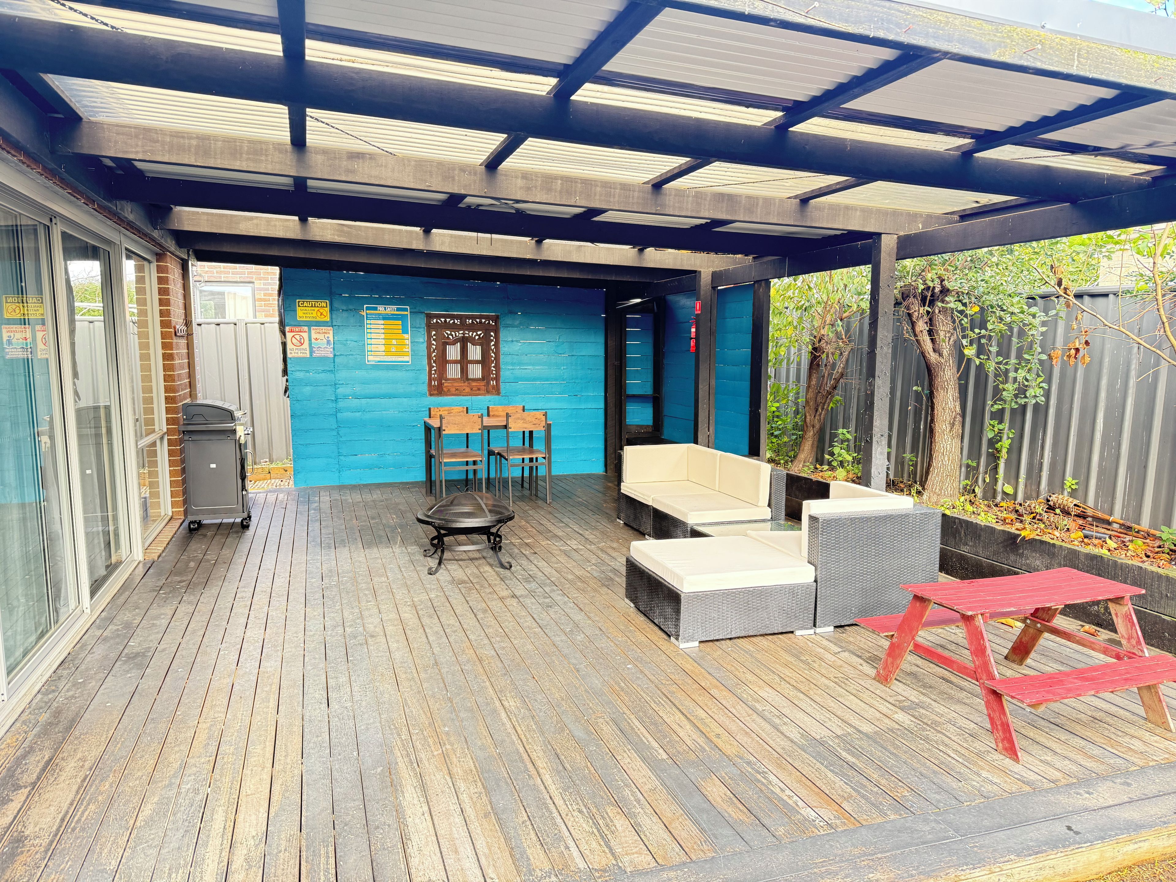 A covered outdoor seating area with a blue wall, wooden chairs, a bench, and a red table.