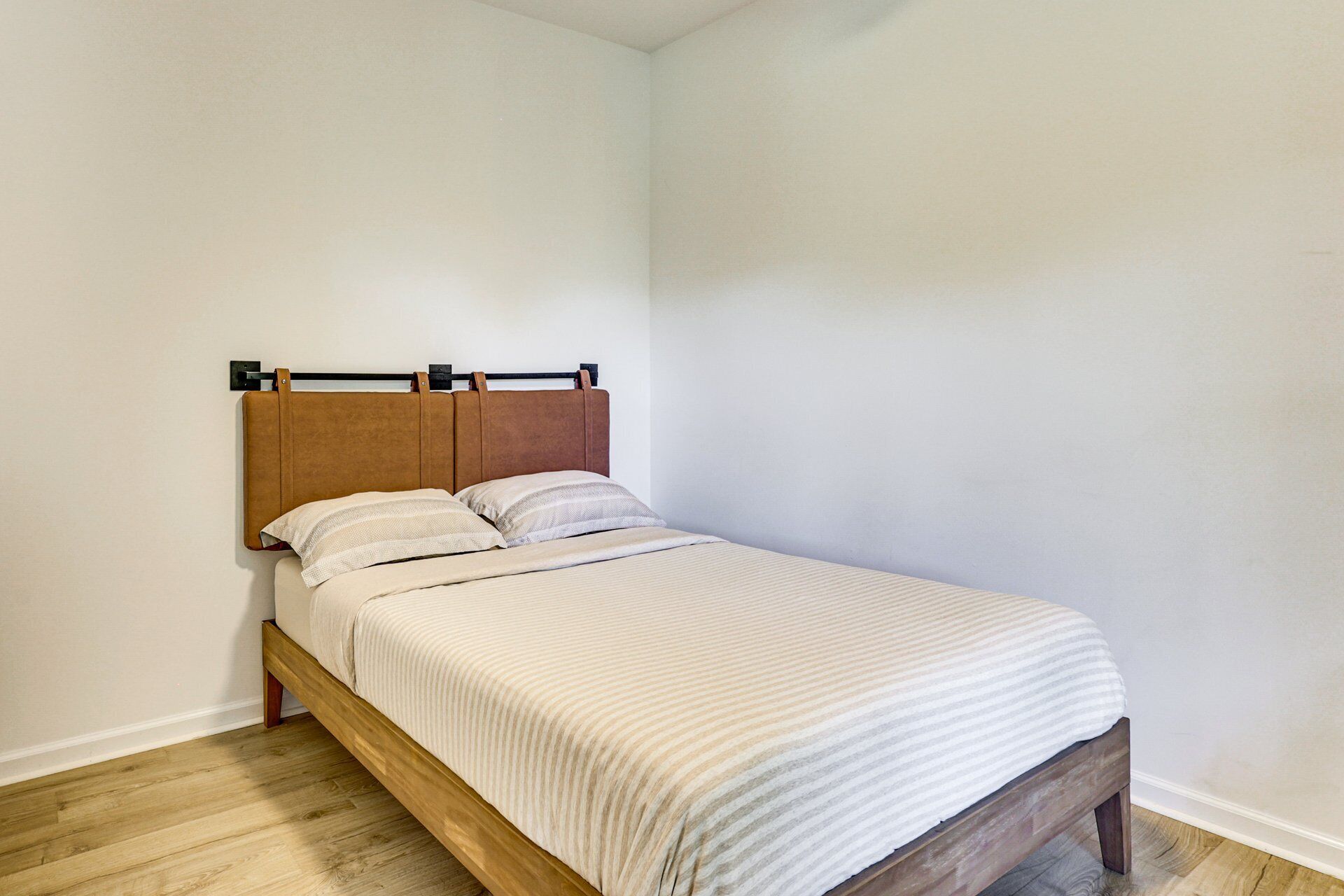 Uma cama de solteiro com estrutura e cabeceira de madeira, uma colcha listrada branca e duas almofadas.