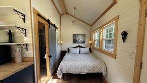 Un dormitorio con cama, piso de madera, ventana y puerta que da a un baño.