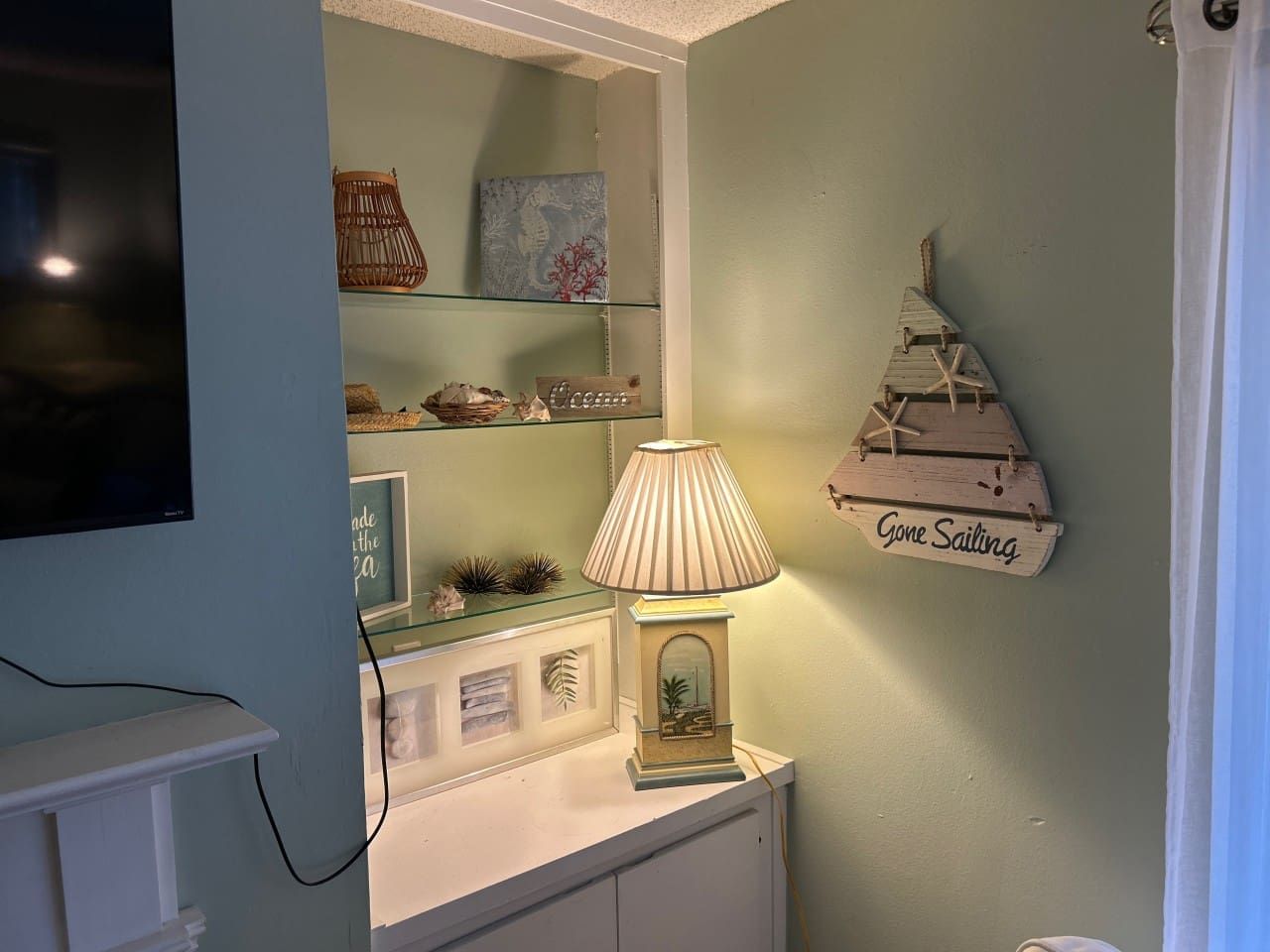 A room with a television mounted on the wall, a lamp on a table, and a shelf with decorative items.