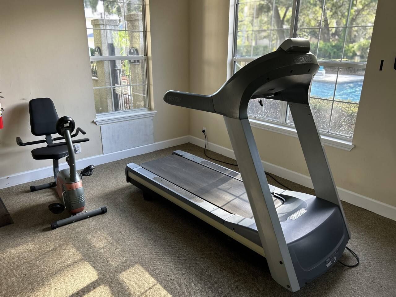 A treadmill in a room with a window, carpeted floor, and a chair.