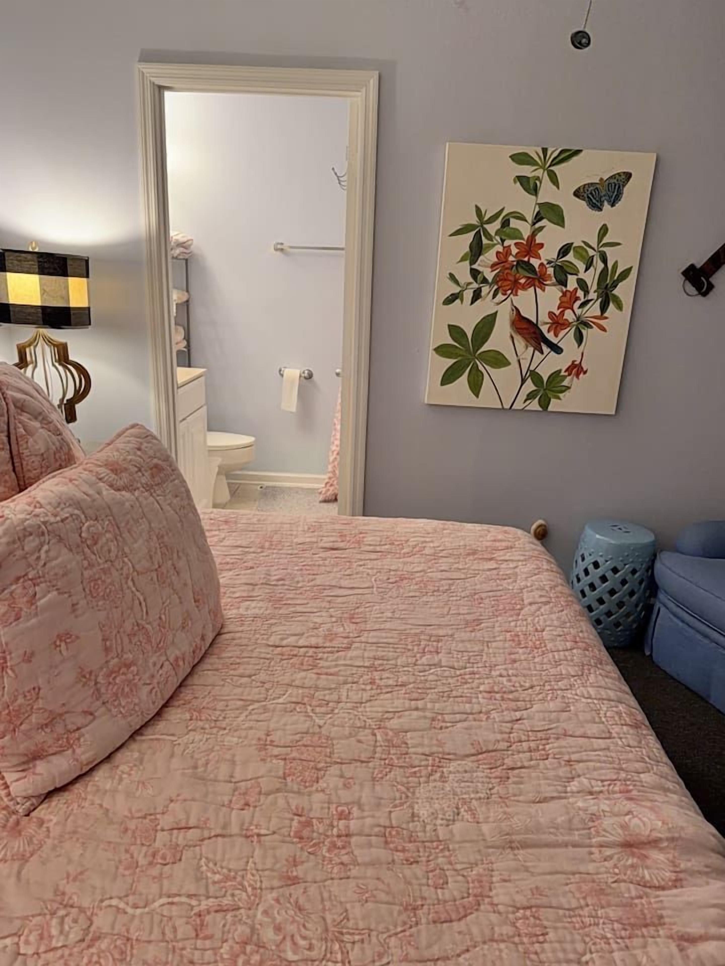 A bedroom with a bed, a blue chair, a painting, and a bathroom visible through an open door.