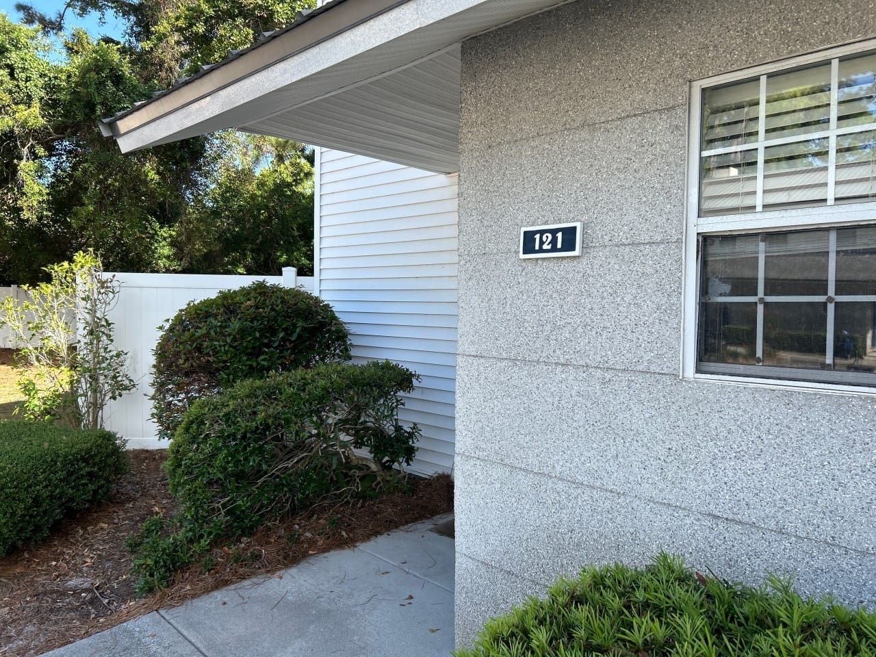 Uma casa com placa de número 121, janela com persianas e um pequeno jardim com arbustos.