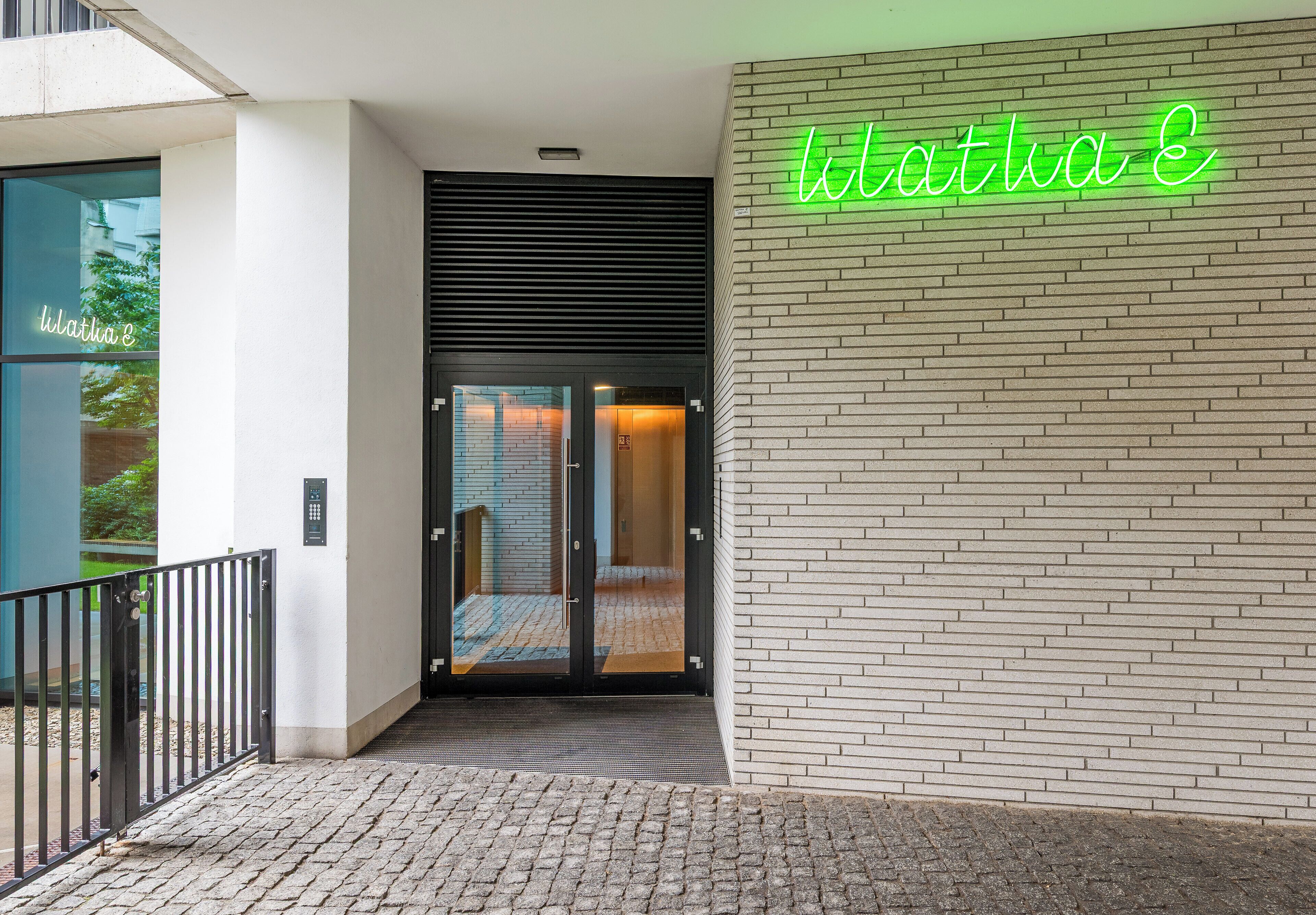 Ein Gebäudeeingang mit einem leuchtenden Neonzeichen mit der Aufschrift 'Kathia G' und einer Glastür, die zu einem Innenbereich führt.