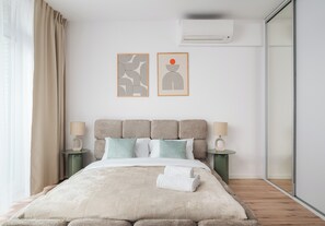 Ein modernes Schlafzimmer mit einem großen Bett, zwei Nachttischen inklusive Lampen, einer Wand-Klimaanlage und zwei abstrakten Wandbildern.