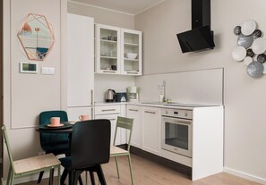 Eine kompakte Küche mit weißen Schränken, einem Backofen, einer Kaffeemaschine und einer Essecke mit zwei Stühlen.