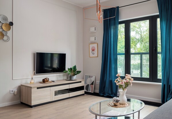 En moderne stue med et fladskærms-tv, et træskab, et glasbord og et vindue med turkise gardiner.