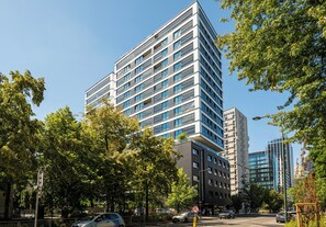 A modern multi-story building with a mix of glass and concrete facades, surrounded by trees and parked cars. - Grzybowska Designer Apartment (Wola)