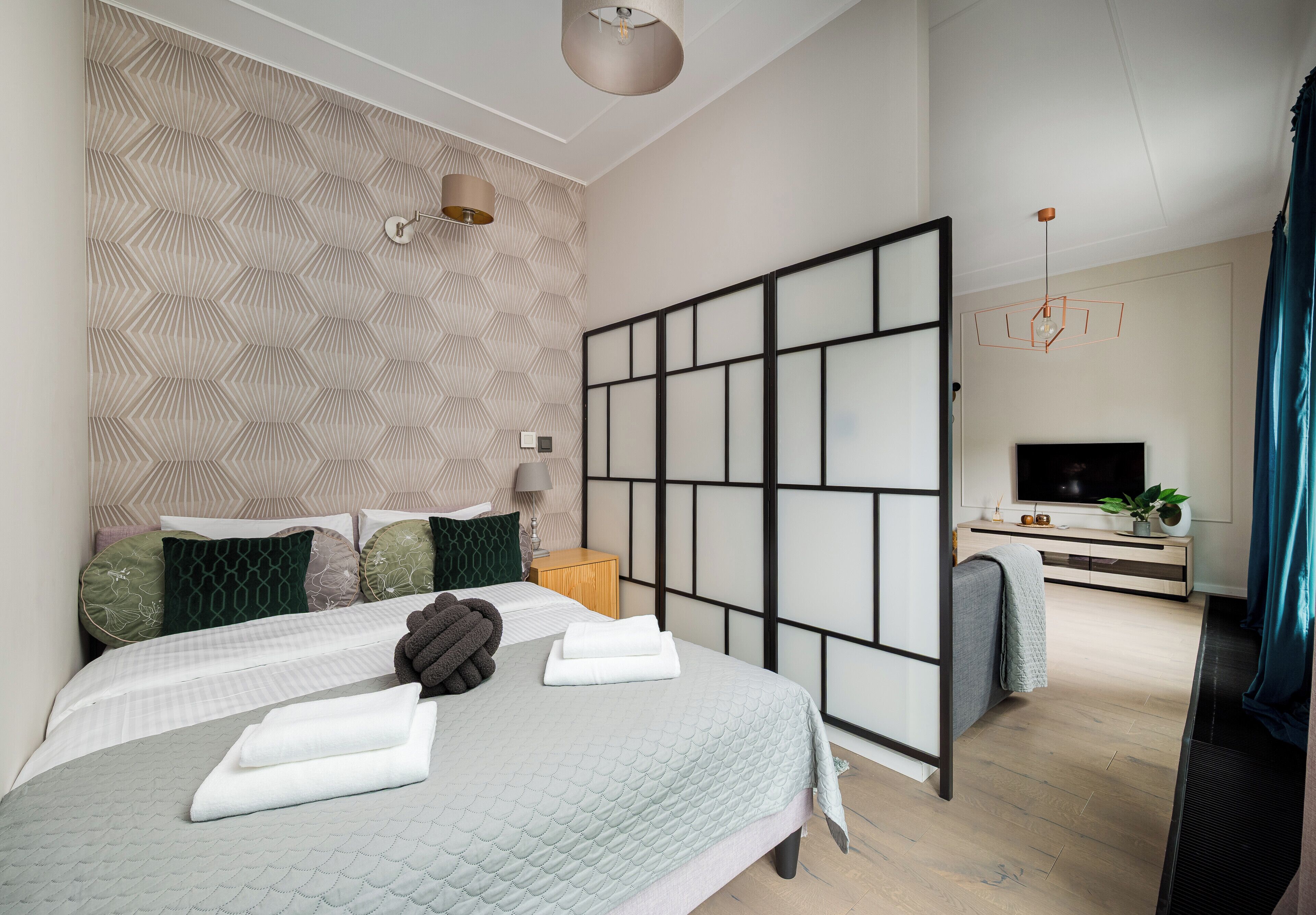 Un dormitorio moderno con cama, almohadas, una mesita de noche, un televisor y una puerta corredera que da a una sala de estar.