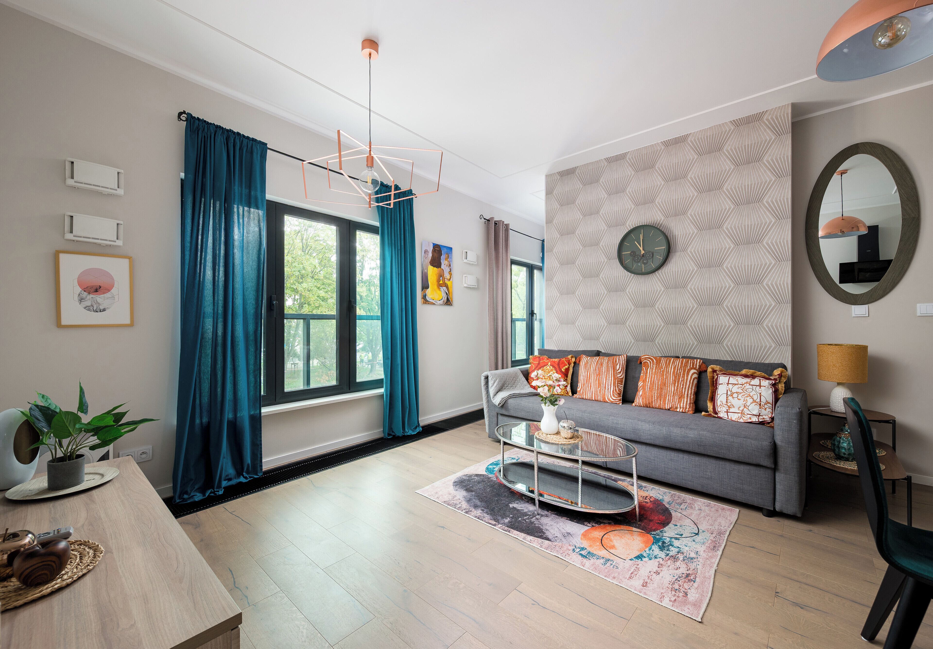 A modern living room with a grey sofa, a coffee table, a large window with curtains, a wall clock, and a mirror.