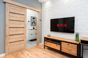 A modern living room with a wooden floor, a TV mounted on a white brick wall, and a wooden cabinet with drawers. - Reymonta 34 Comfort Spot Studio (Katowice)