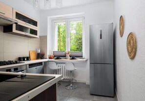 A modern kitchen with a stainless steel refrigerator, a built-in oven, and a window overlooking greenery. - Reymonta 34 Comfort Spot Studio (Katowice)