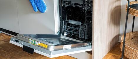 Un lavavajillas abierto con un guante de cocina azul colgando en la pared, una silla de mimbre y un suelo de madera.