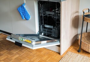 An open dishwasher with a blue oven mitt hanging on the wall, a wicker chair, and a wooden floor. - Solec Powiśle Harmony Studio (Warszawa)