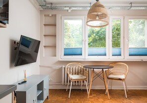 A room with a television mounted on the wall, a table with two wicker chairs, a hanging light fixture, and a window with blinds. - Solec Powiśle Harmony Studio (Warszawa)