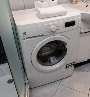 Edestä täytettävä Electrolux-pesukone, jossa on ohjauspaneeli, päällä valkoinen pyyhe ja lähellä wc-paperirulla.