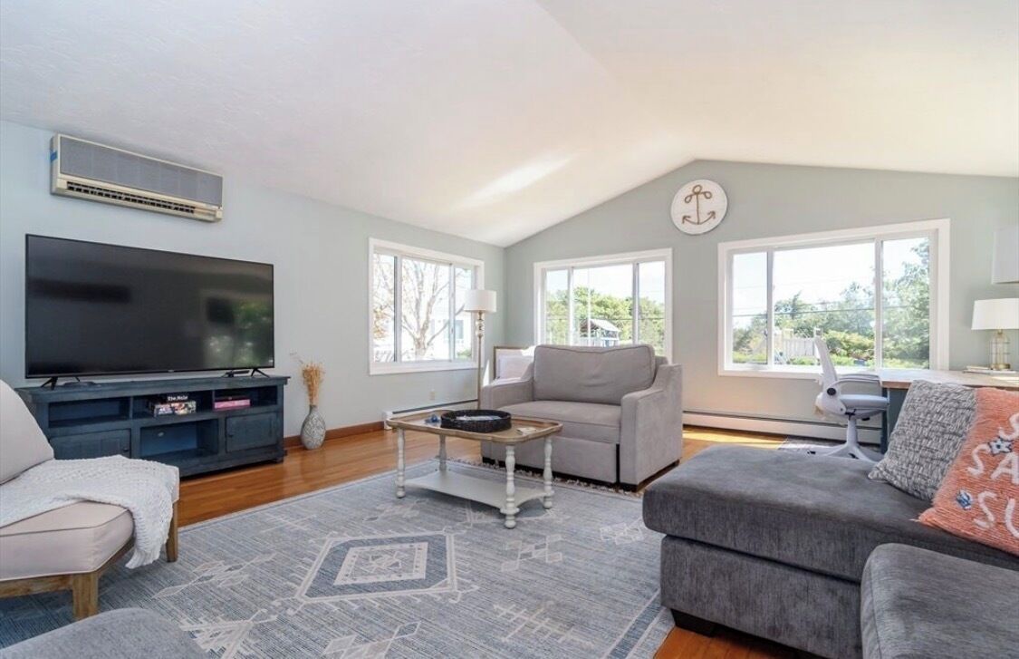 A living room with a flat-screen TV, grey sofas, a coffee table, and large windows.