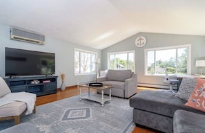 A living room with a flat-screen TV, grey sofas, a coffee table, and large windows.