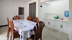 A dining area with a table set for six, wooden chairs, a white tablecloth, a grey curtain, and a kitchen area with cabinets and a water dispenser. - The ritz villa Nuwaraeliya (Nuwara Eliya)