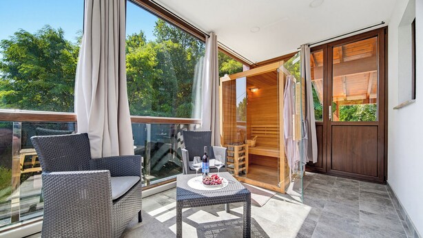 A balcony with wicker furniture, a glass table with a bottle and glass, and a wooden door leading to a sauna. - Greenwood Retreat With Hot Tub & Sauna, Sveti Martin na Muri, Croatia (Sveti Martin na Muri)