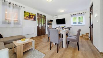 A modern living room with a sofa, a dining table with chairs, a TV mounted on the wall, and a kitchen area with a window.