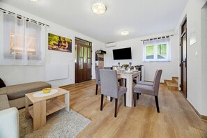 A modern living room with a sofa, a dining table with chairs, a TV mounted on the wall, and a kitchen area with a window. - Greenwood Retreat With Hot Tub & Sauna, Sveti Martin na Muri, Croatia (Sveti Martin na Muri)