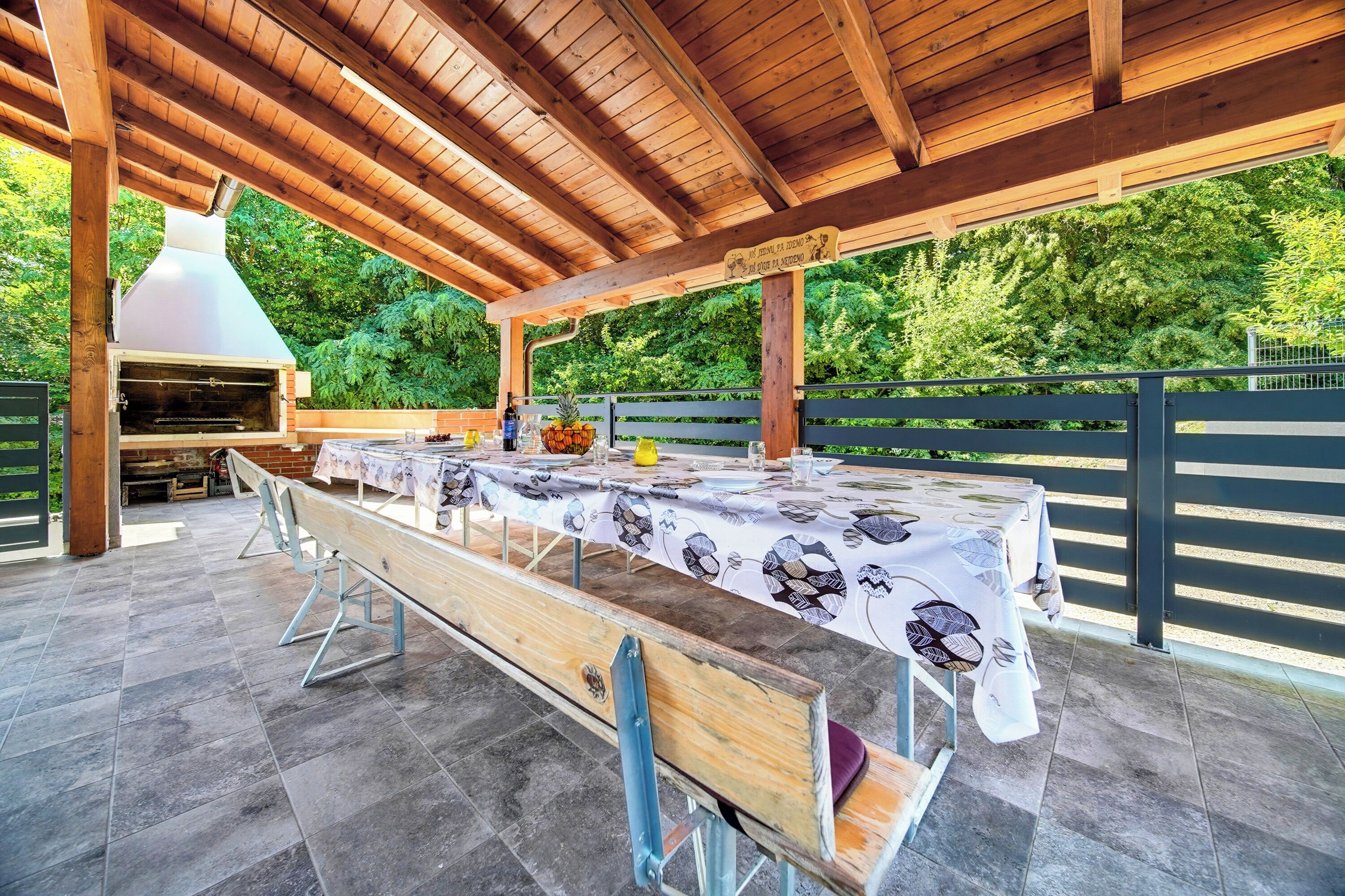 An outdoor patio with a long wooden table set for dining, a barbecue grill, and a pergola overhead.