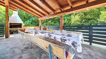 An outdoor patio with a long wooden table set for dining, a barbecue grill, and a pergola overhead.