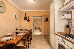 A dining area with a wooden table set for four, a wine bottle, and a bowl of fruit. There is a stone wall in the background and a kitchen with white cabinets and a stove. - Villa Dalmatia | Luxury Stone Villa with Pool & Tennis, Biograd na Moru, Croatia (Biograd na Moru)