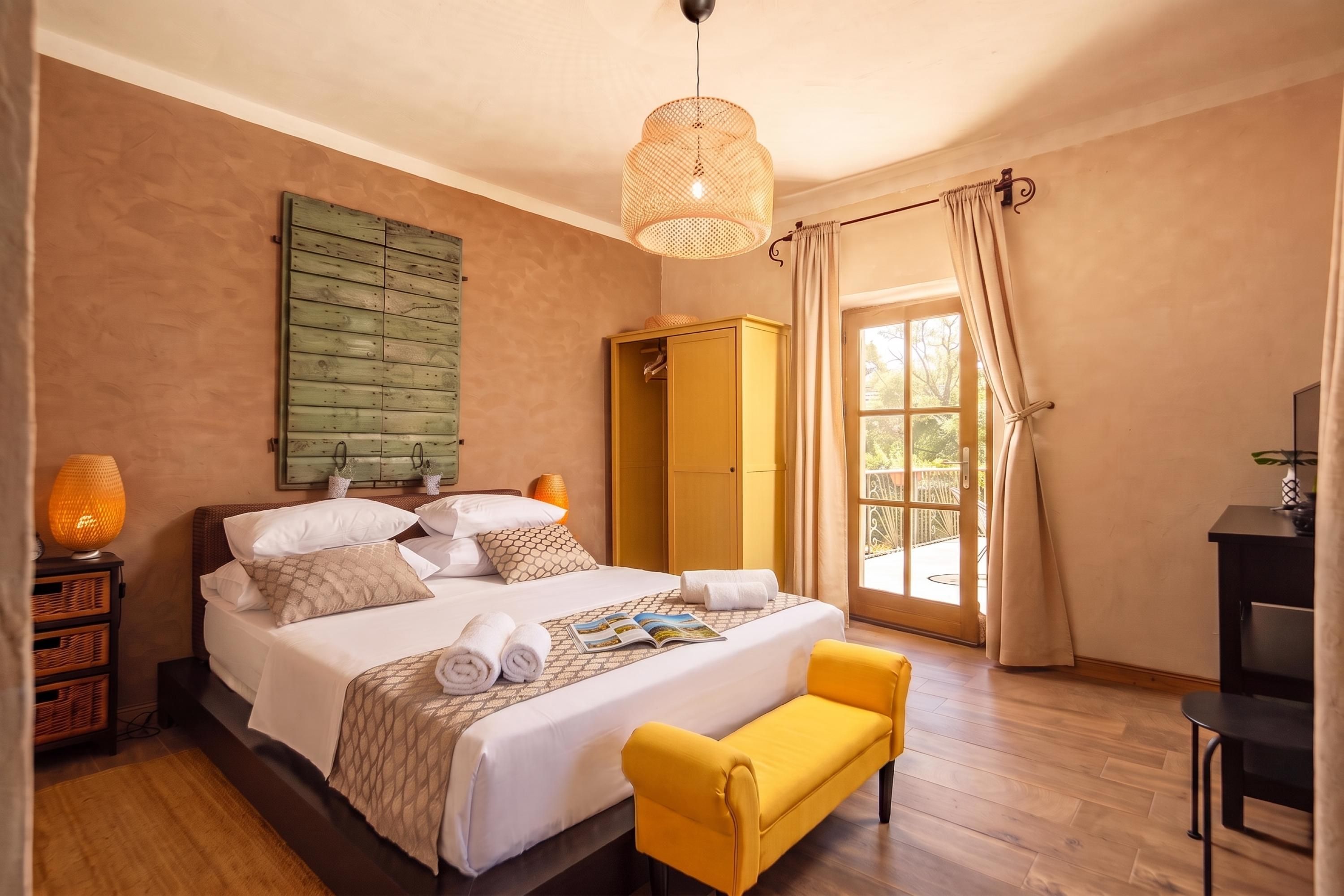 A hotel room with a bed, a yellow armchair, a wardrobe, and a large window with curtains.