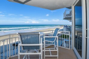 Ein Balkon mit Blick auf Strand und Meer, weiße Gartenmöbel und eine Glastür.