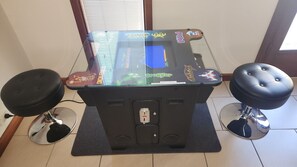 Ein Retro-Spielautomat mit Glasabdeckung, zwei Hocker und einer Bedieneinheit.