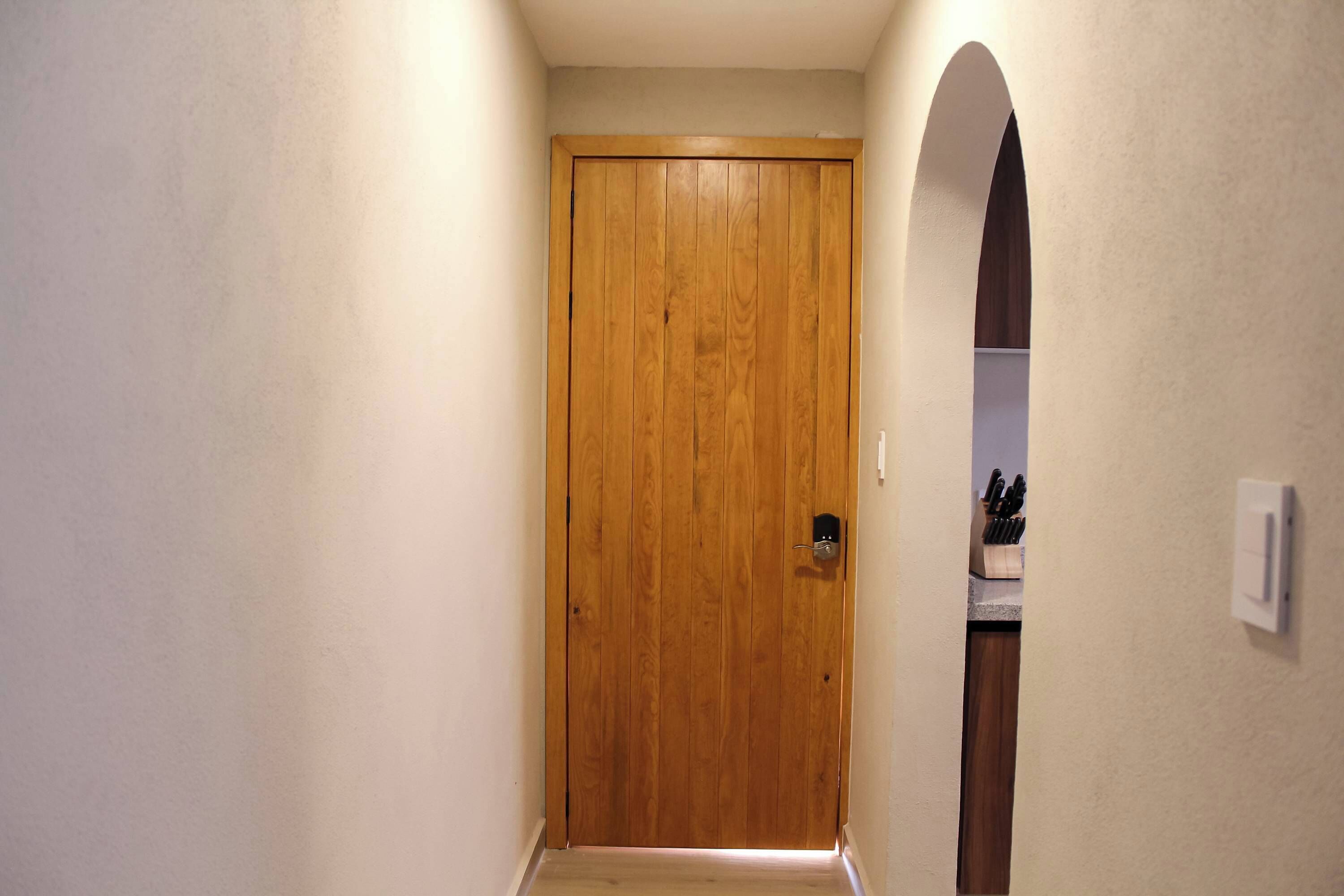 A wooden door in a hallway with a light switch on the wall and a knife block in the background.