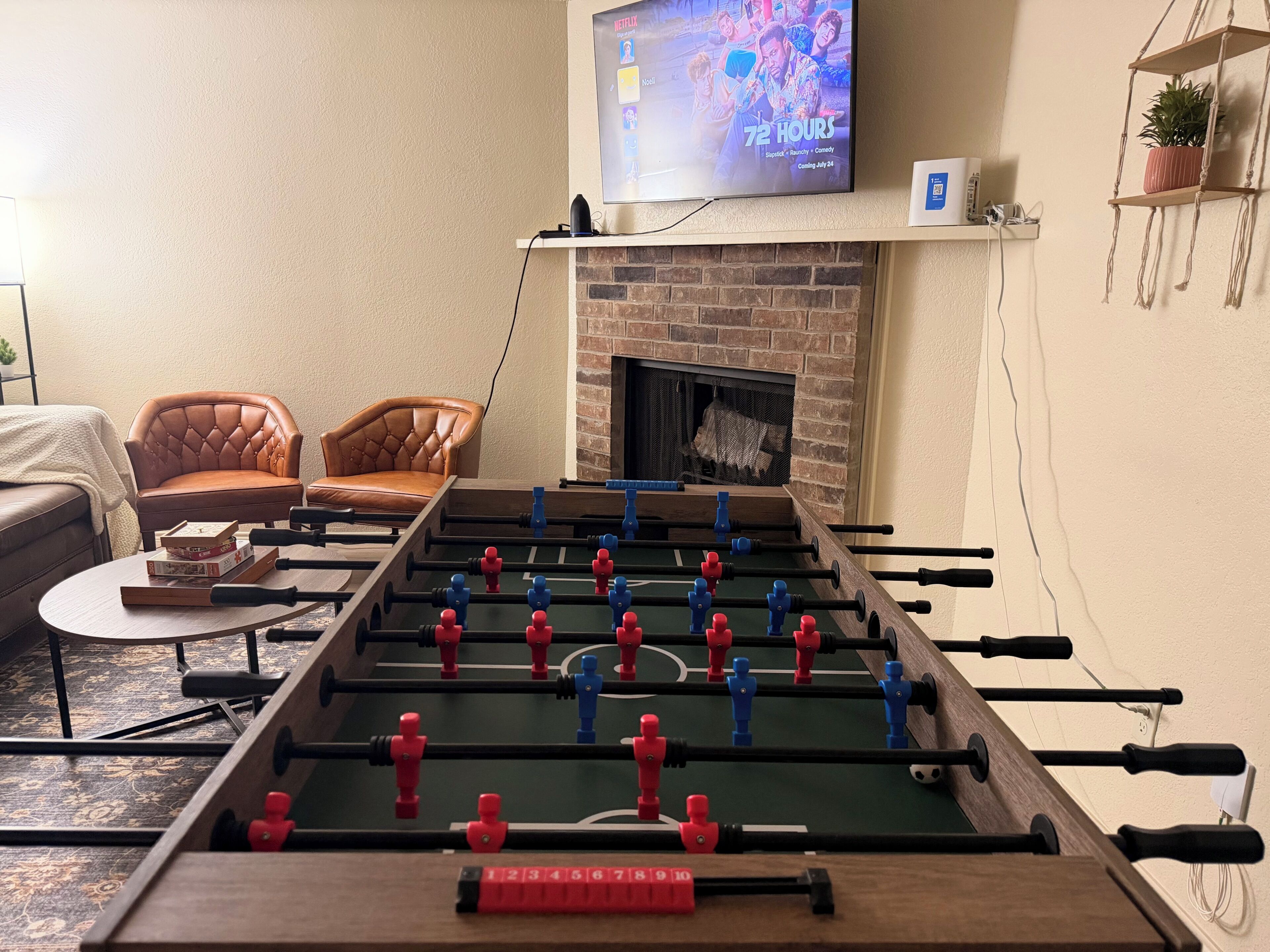 Una sala de estar con una mesa de fútbol, un sofá, un televisor sobre la chimenea y un estante pequeño con una planta.