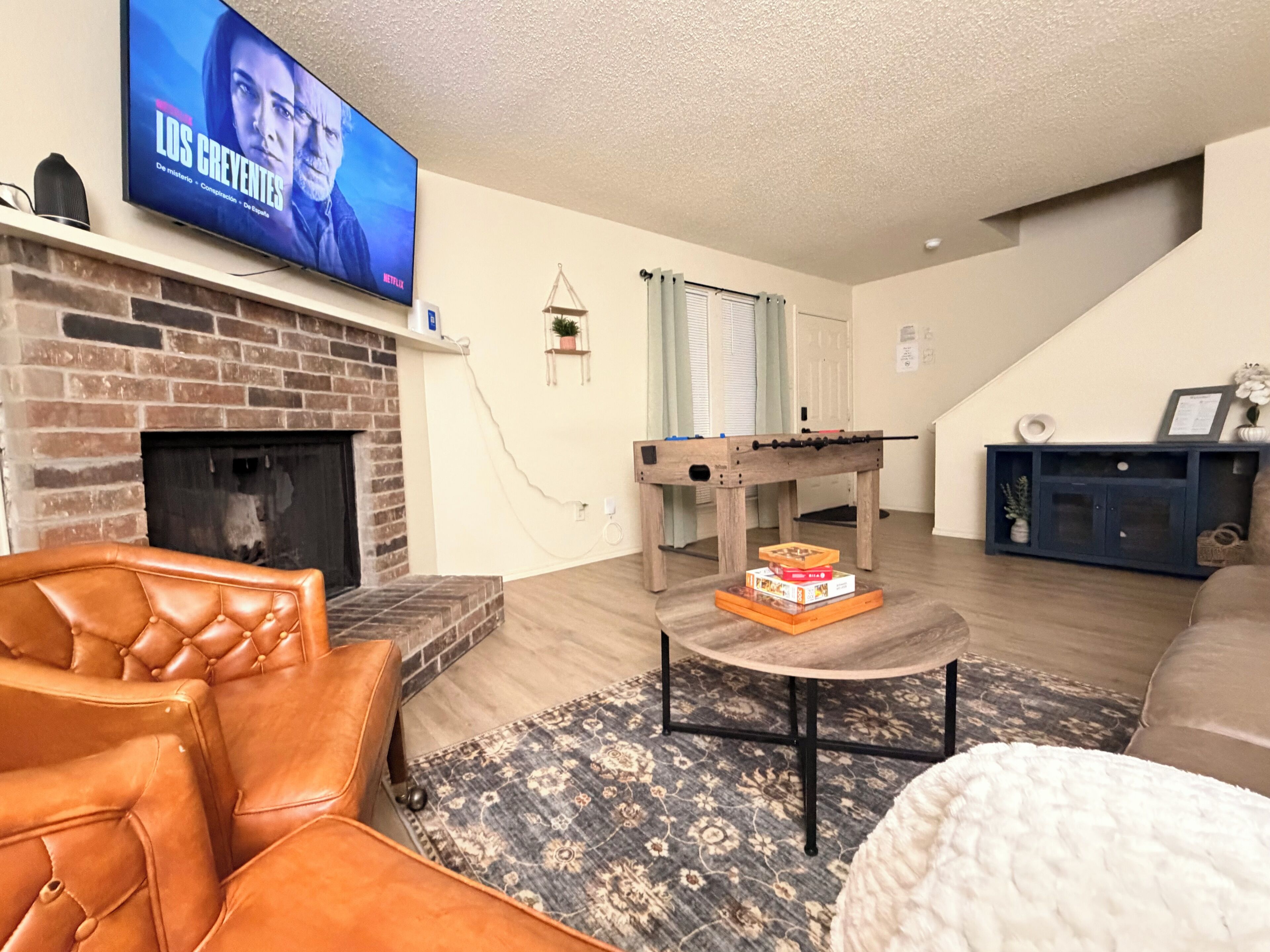A living room with a leather sofa, a fireplace, a foosball table, and a television displaying a show.