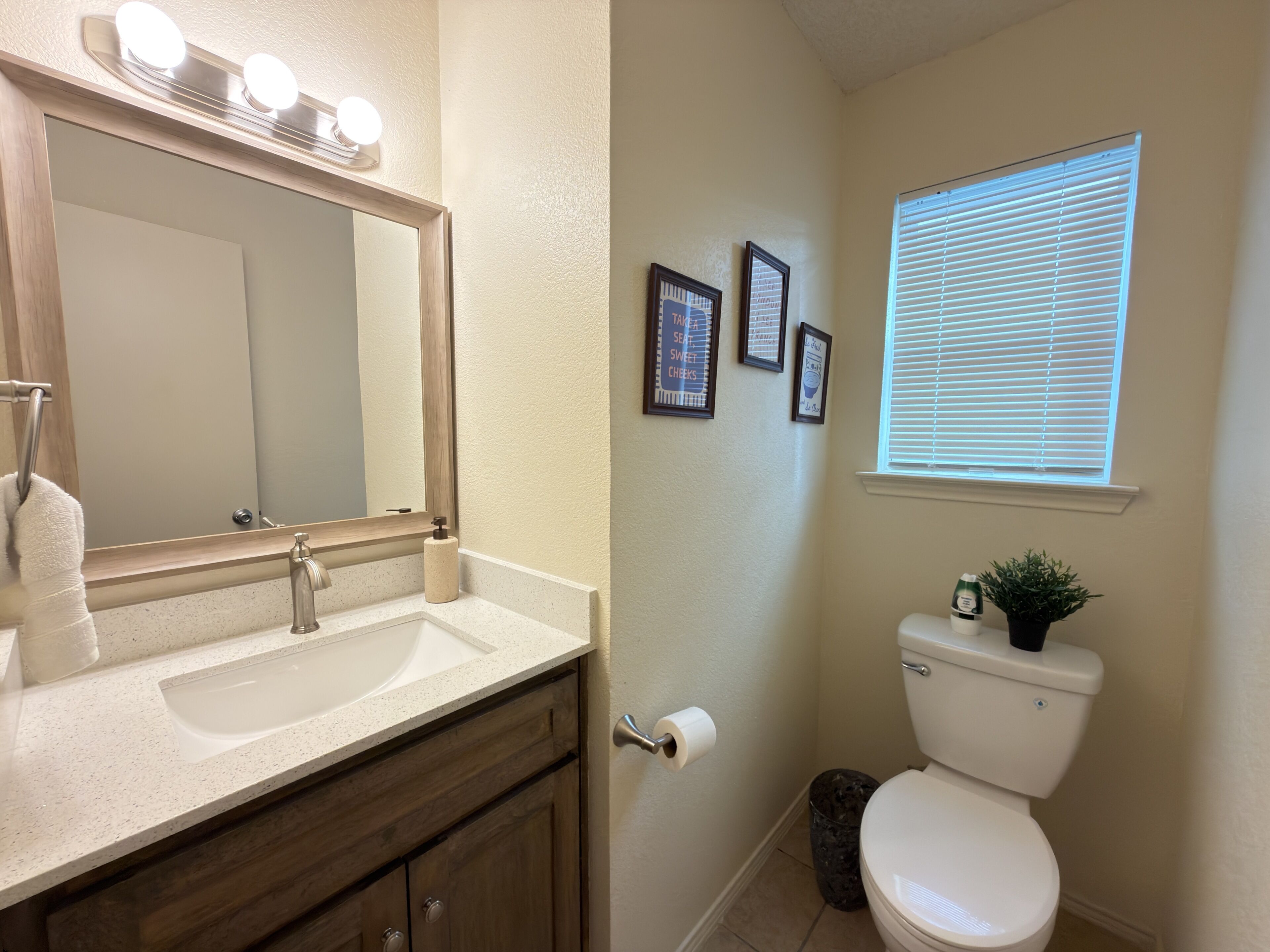 A bathroom with a double sink vanity, a toilet, a window with blinds, and a mirror with lights above.