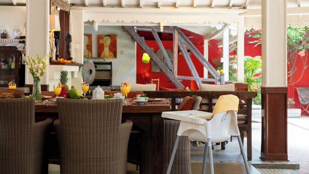 Un coin repas avec des meubles en osier, une chaise blanche et une table dressée avec des fruits, des boissons et une carafe.