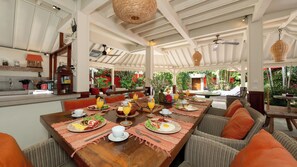 A dining area with a long wooden table set for breakfast, surrounded by wicker chairs, and a kitchen area with a sink and cabinets. - Villa Hangat by Villa Finder (Badung)