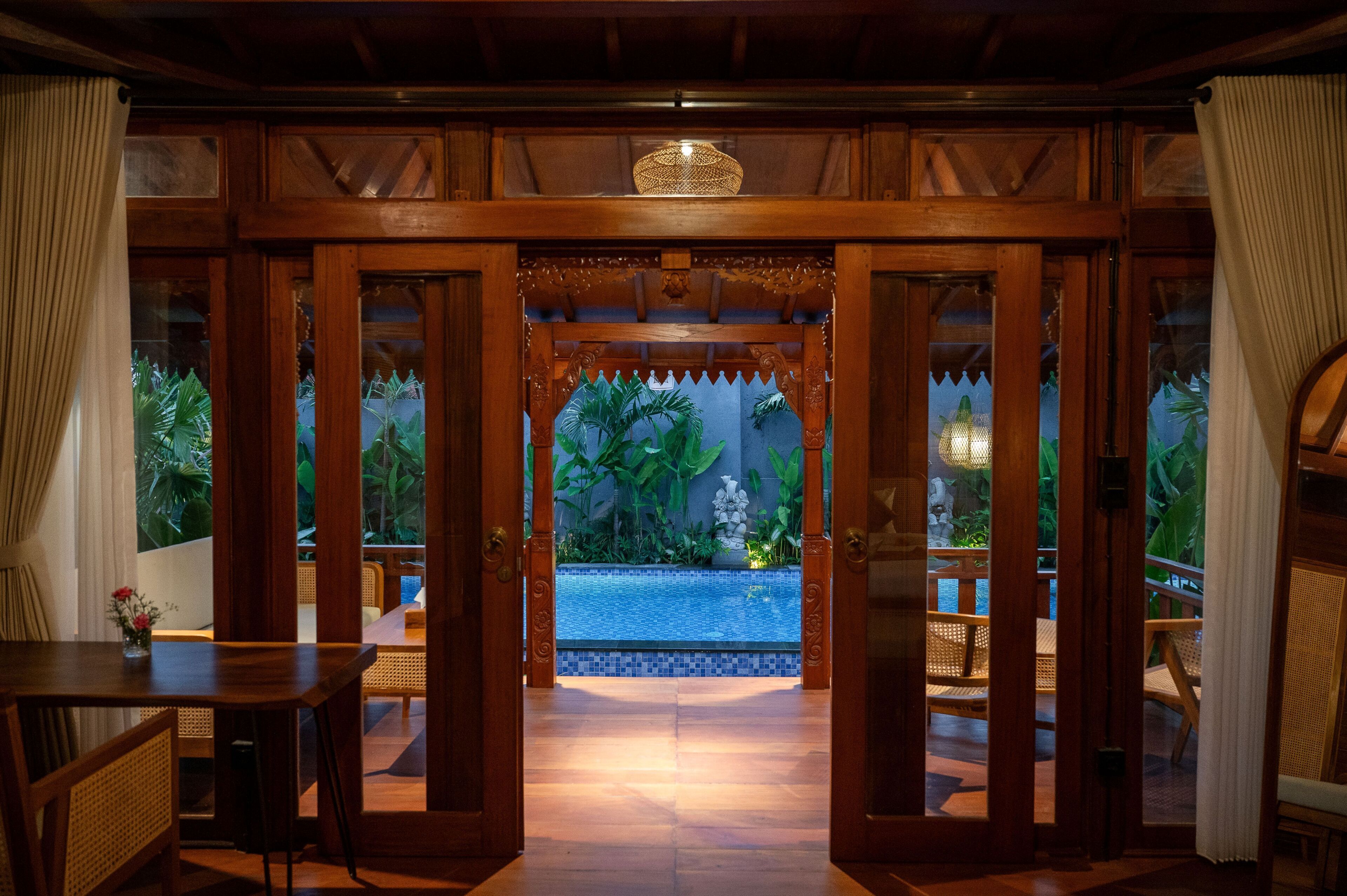 Puerta de madera abierta que da a una zona de piscina con suelo de baldosas y vistas a plantas tropicales.