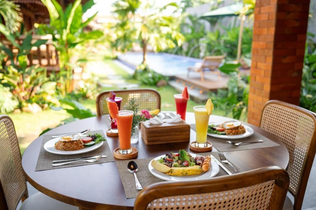 A round dining table with plates of food, drinks, and a small wooden box, set in an outdoor garden with a pool in the background.