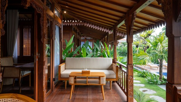 Un porche de madera con una zona de estar acolchada, una mesita y vistas a un jardín con plantas tropicales.