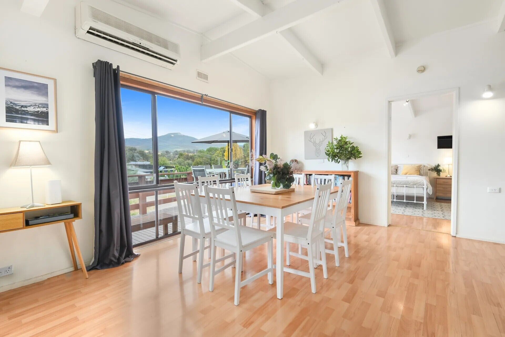 A spacious room with a wooden dining table, white chairs, a large window with a view of mountains, and an air conditioning unit.